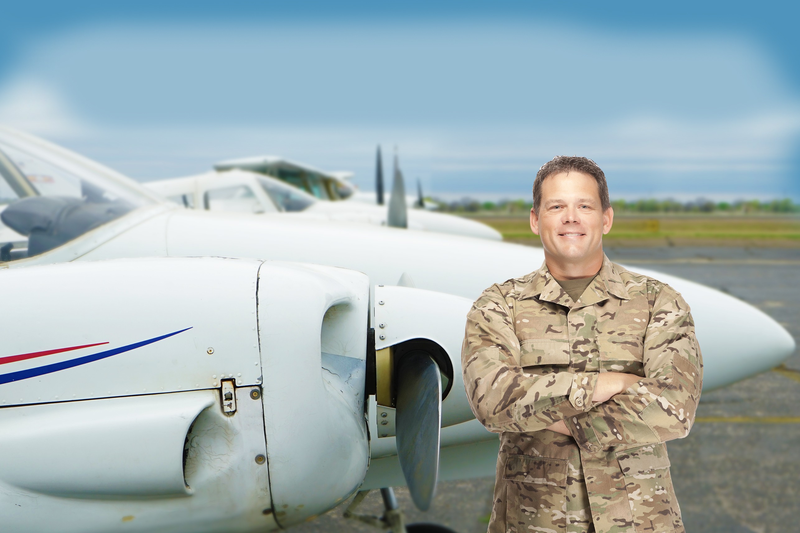 Smiling male military pilot or service member in camouflage uniform standing with arms crossed in front of a twin-engine propeller aircraft.