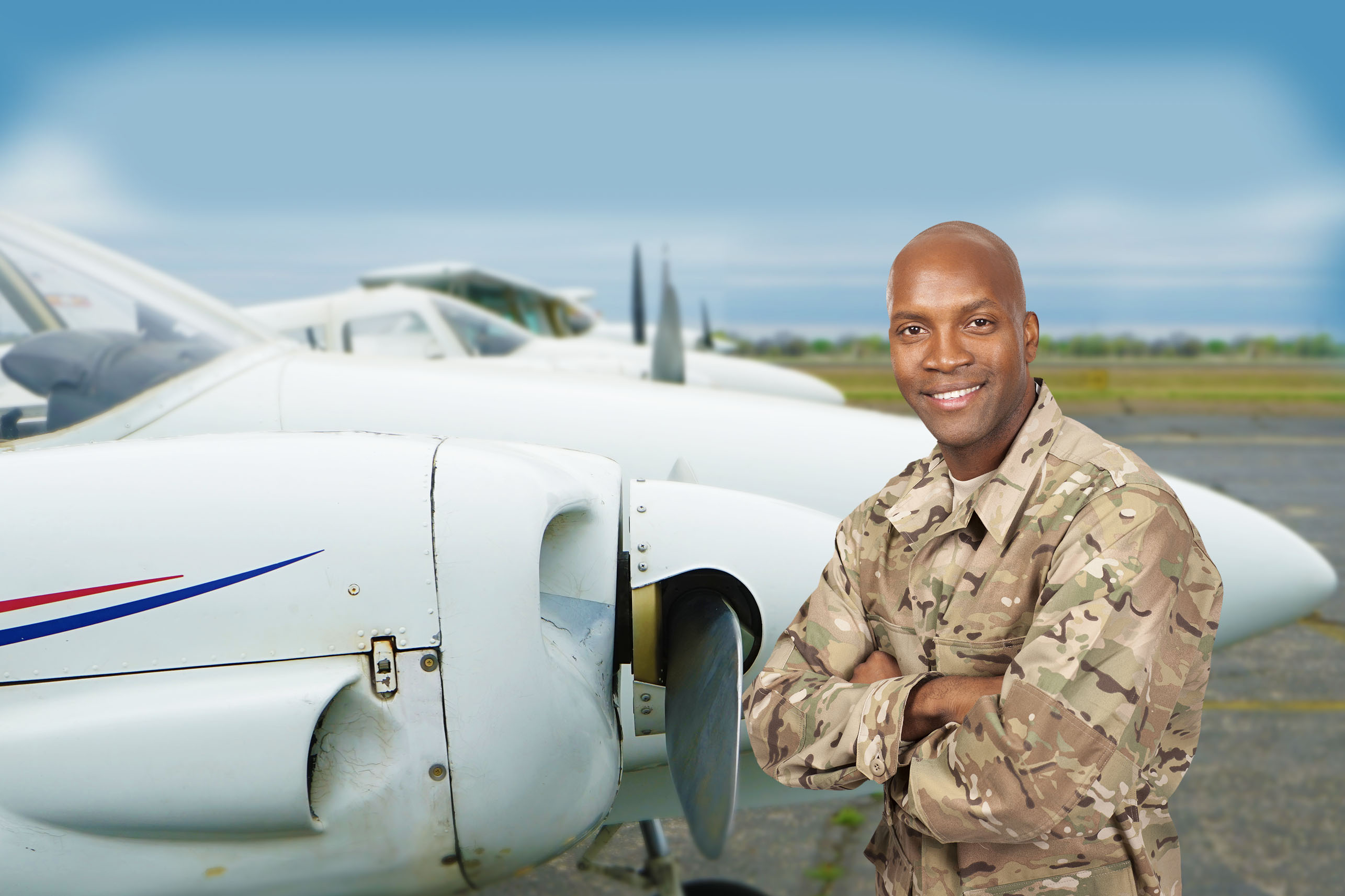 Smiling Black male pilot or soldier in military camouflage uniform standing with arms crossed in front of a twin-engine aircraft