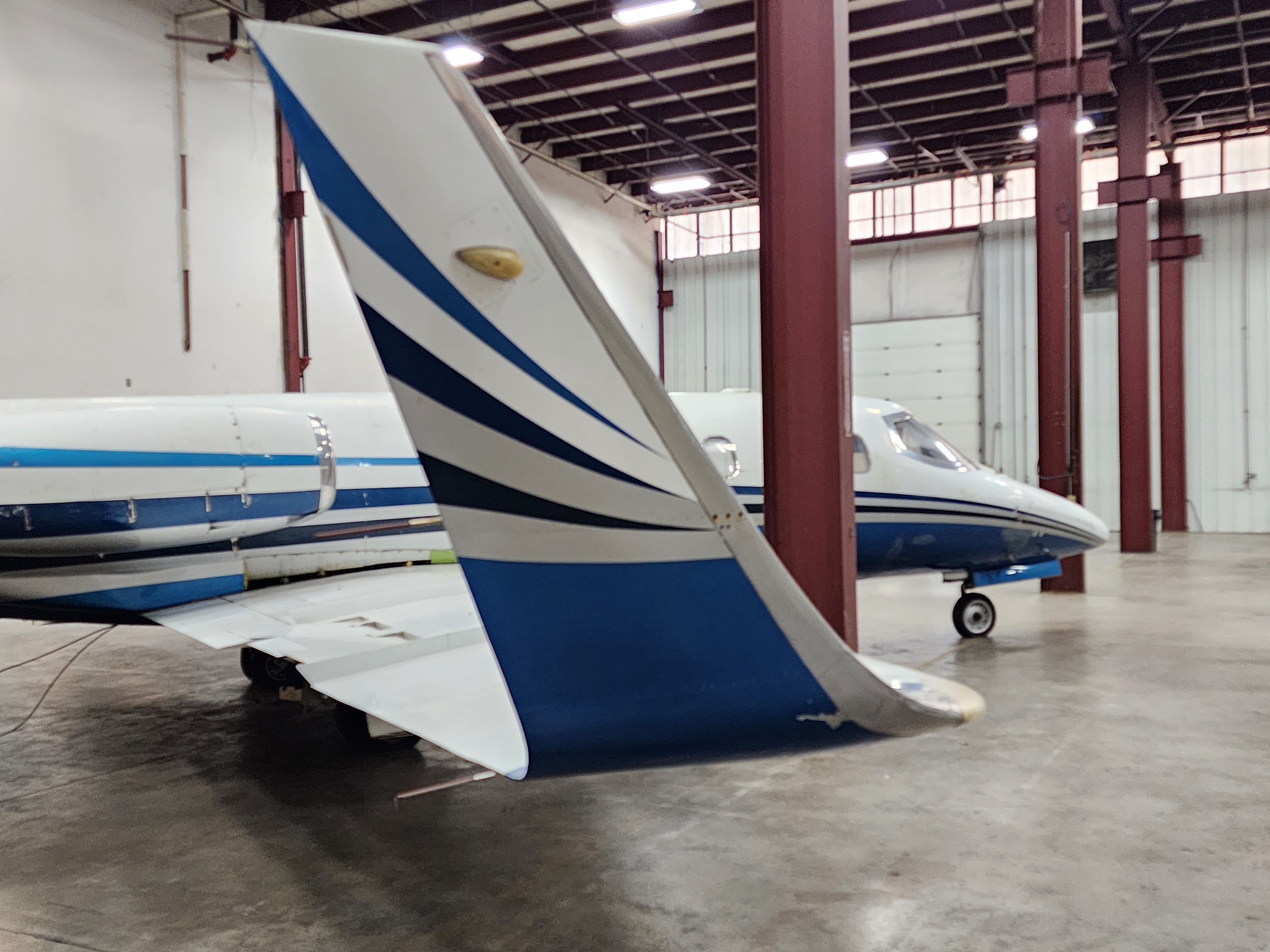 Private jet parked inside a hangar, showing a close-up of the tail section with blue and white striped detailing.