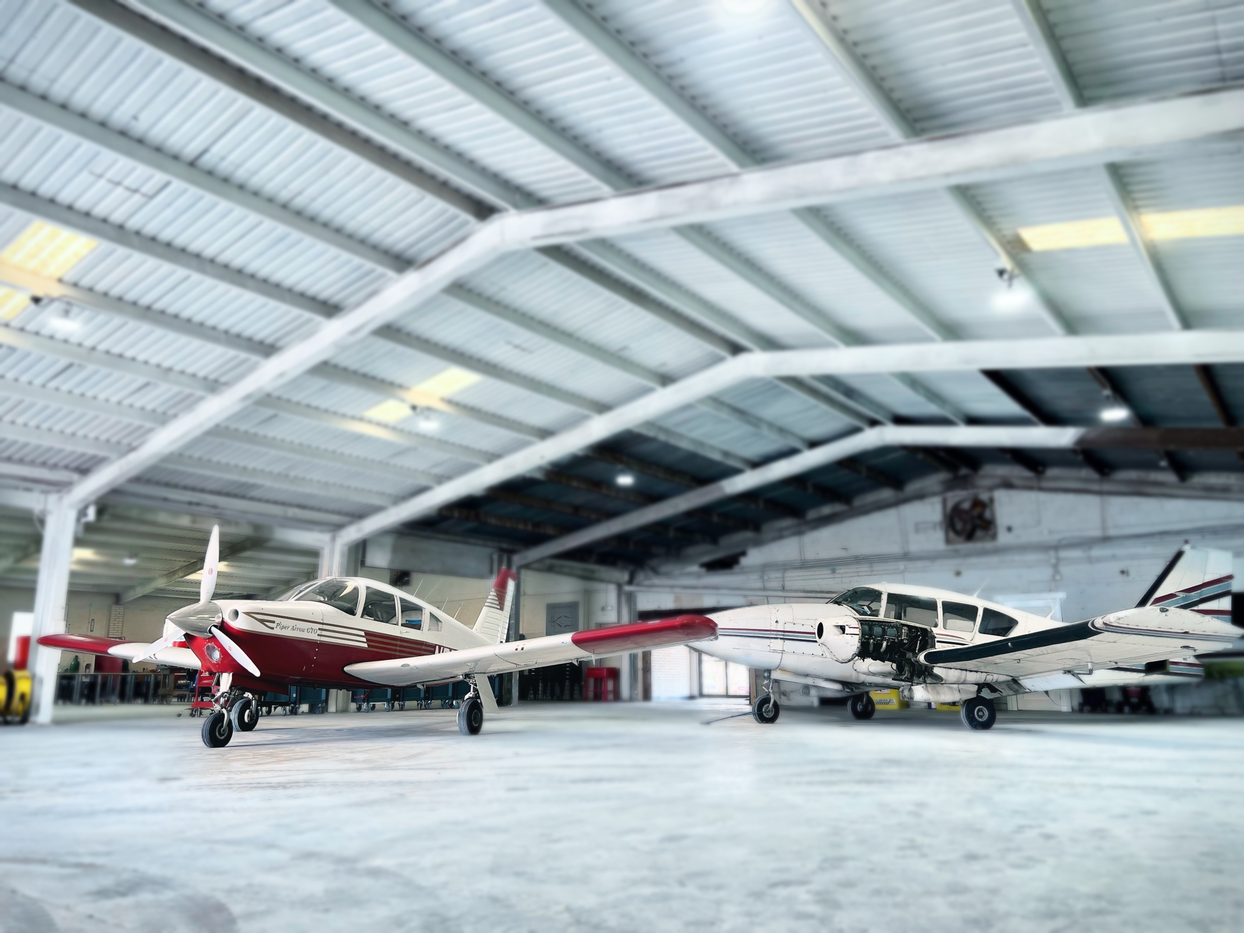 A pair of small twin-engine training aircraft parked inside a hangar, with one plane’s engine cowling removed for maintenance.