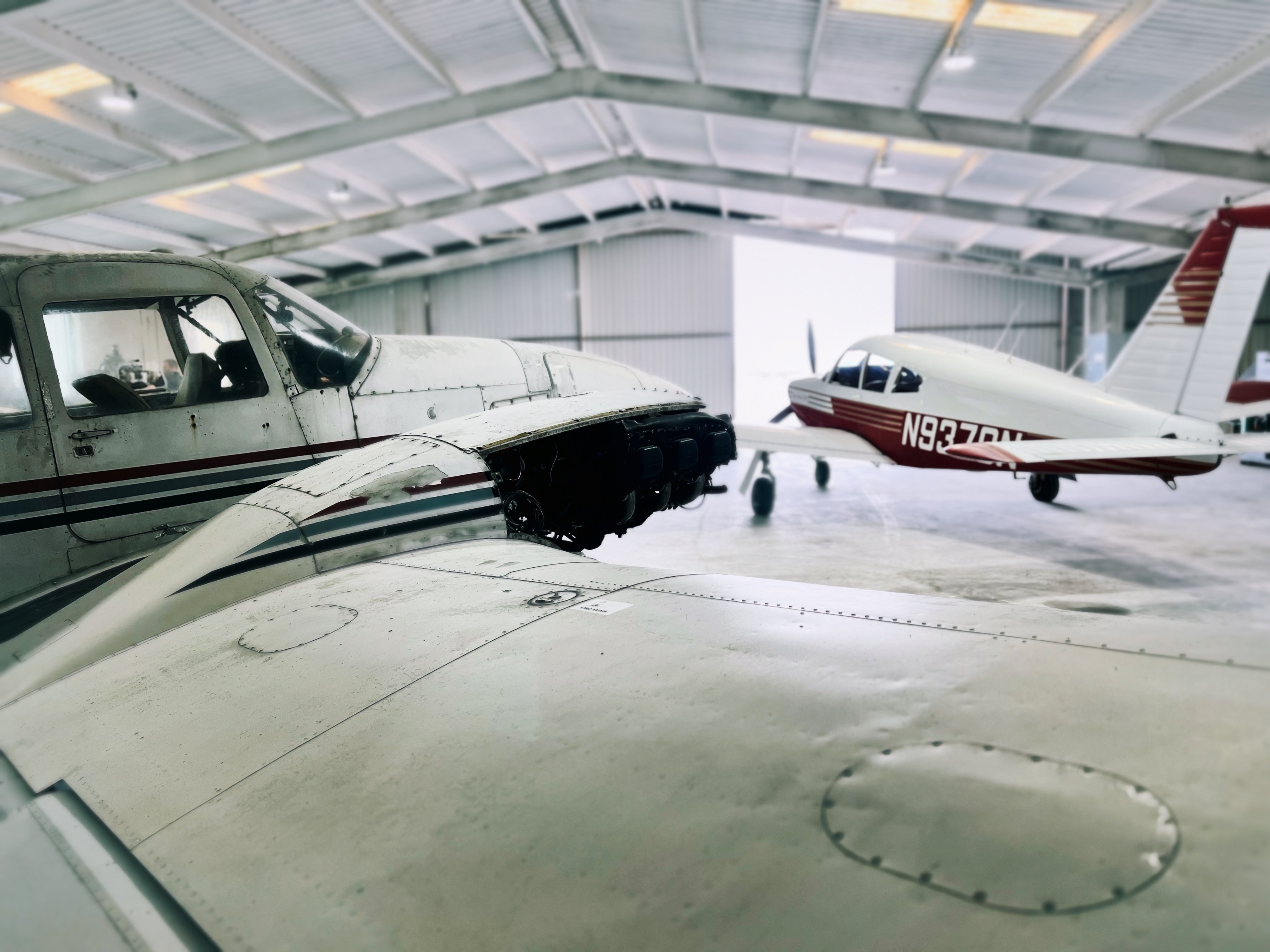 A partially disassembled small aircraft inside a hangar, showing an exposed engine on the wing with another parked airplane in the background.