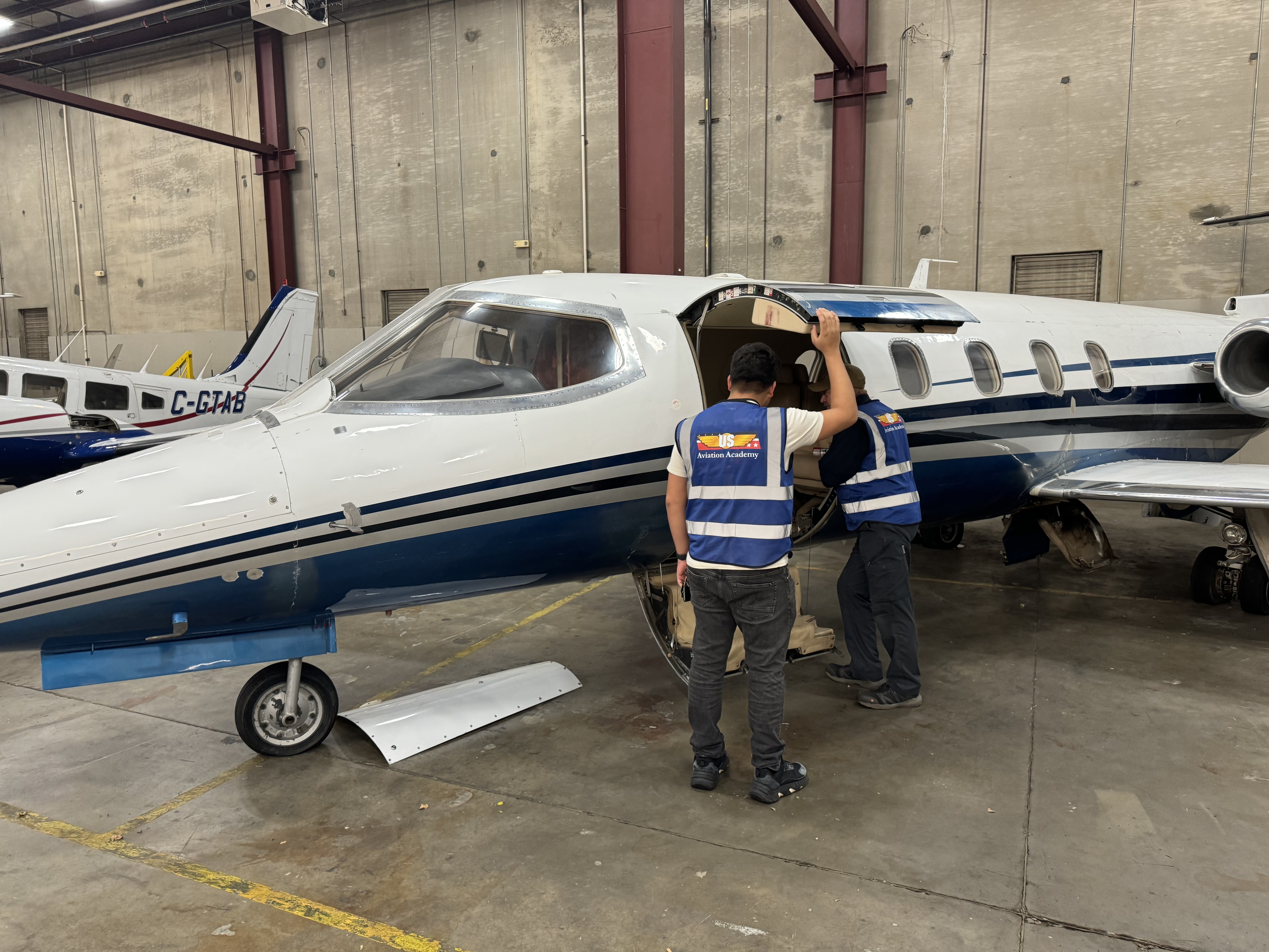 Two aviation students in safety vests inspecting the interior of a small jet inside a maintenance hangar.