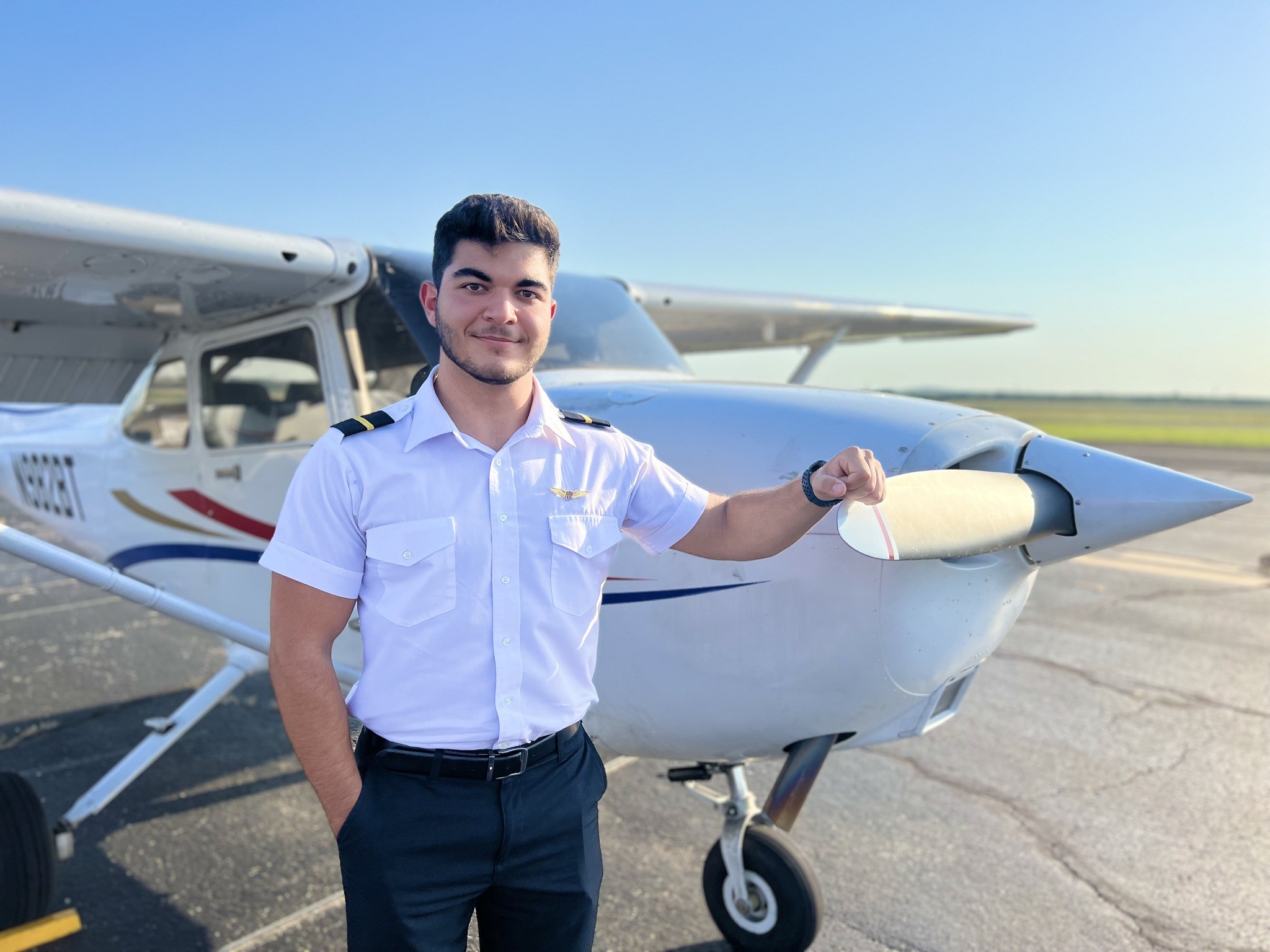 Confident student pilot wearing a uniform with two-stripe epaulets, posing next to the propeller of a single-engine training aircraft at golden hour