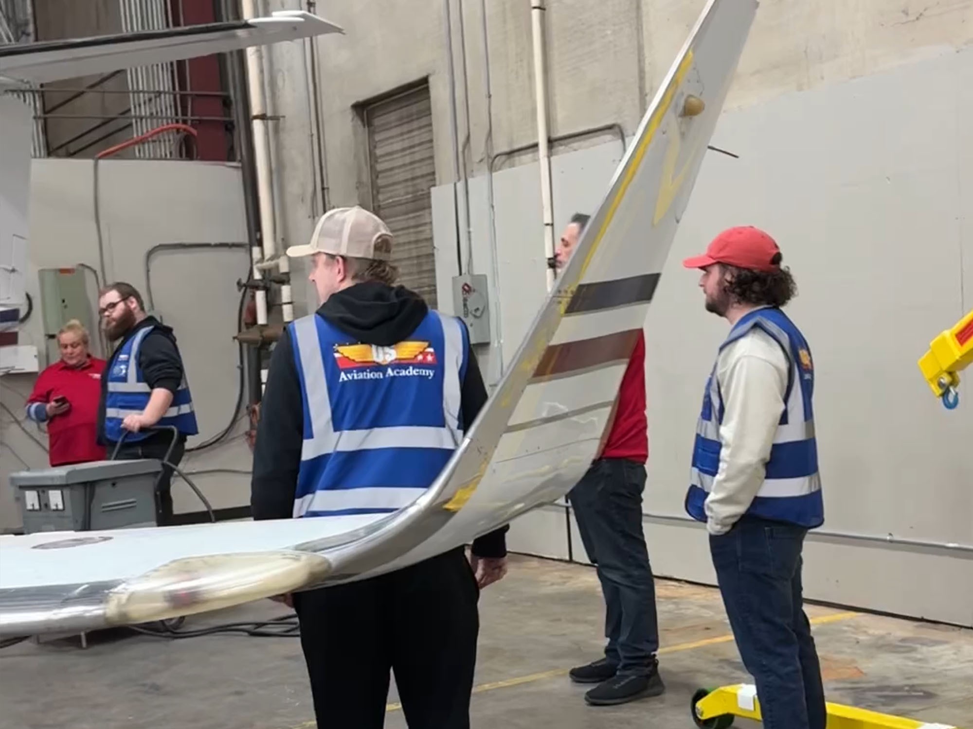 Aviation Academy students in blue safety vests standing beside an aircraft wing inside a maintenance hangar.