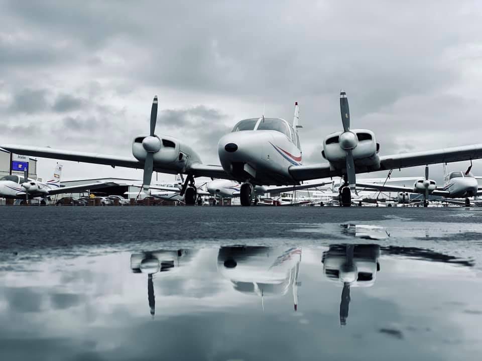 twin-engine propeller aircraft parked on a wet tarmac, reflected in a puddle under a cloudy, overcast sky