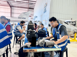 Two aviation maintenance students wearing safety vests and goggles inspecting components on a large aircraft engine during hands-on training in a hangar.