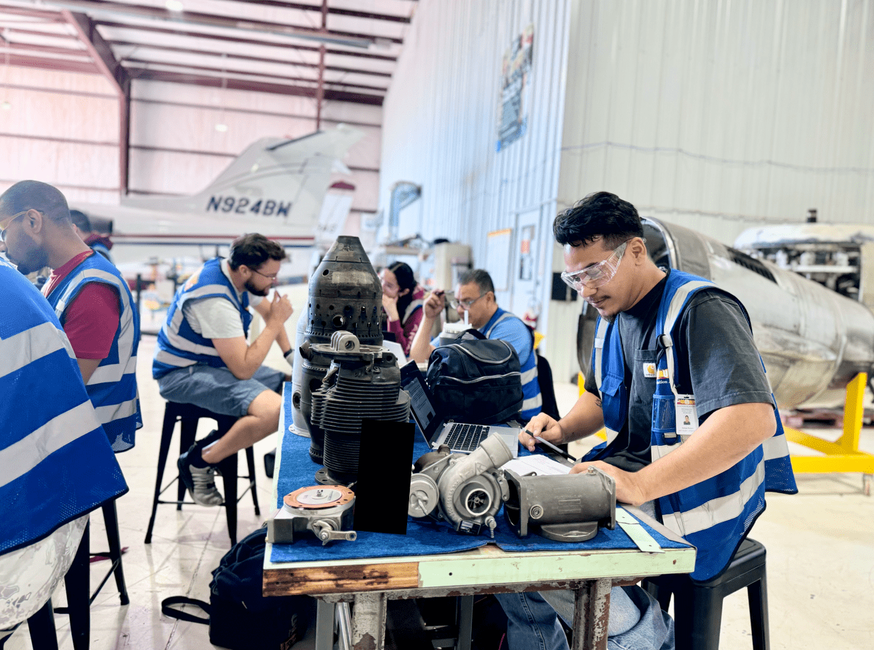 Two aviation maintenance students wearing safety vests and goggles inspecting components on a large aircraft engine during hands-on training in a hangar.