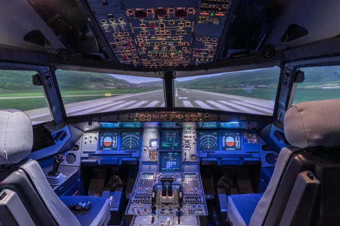 Airline cockpit simulator with illuminated control panels and a runway displayed on the front screens.