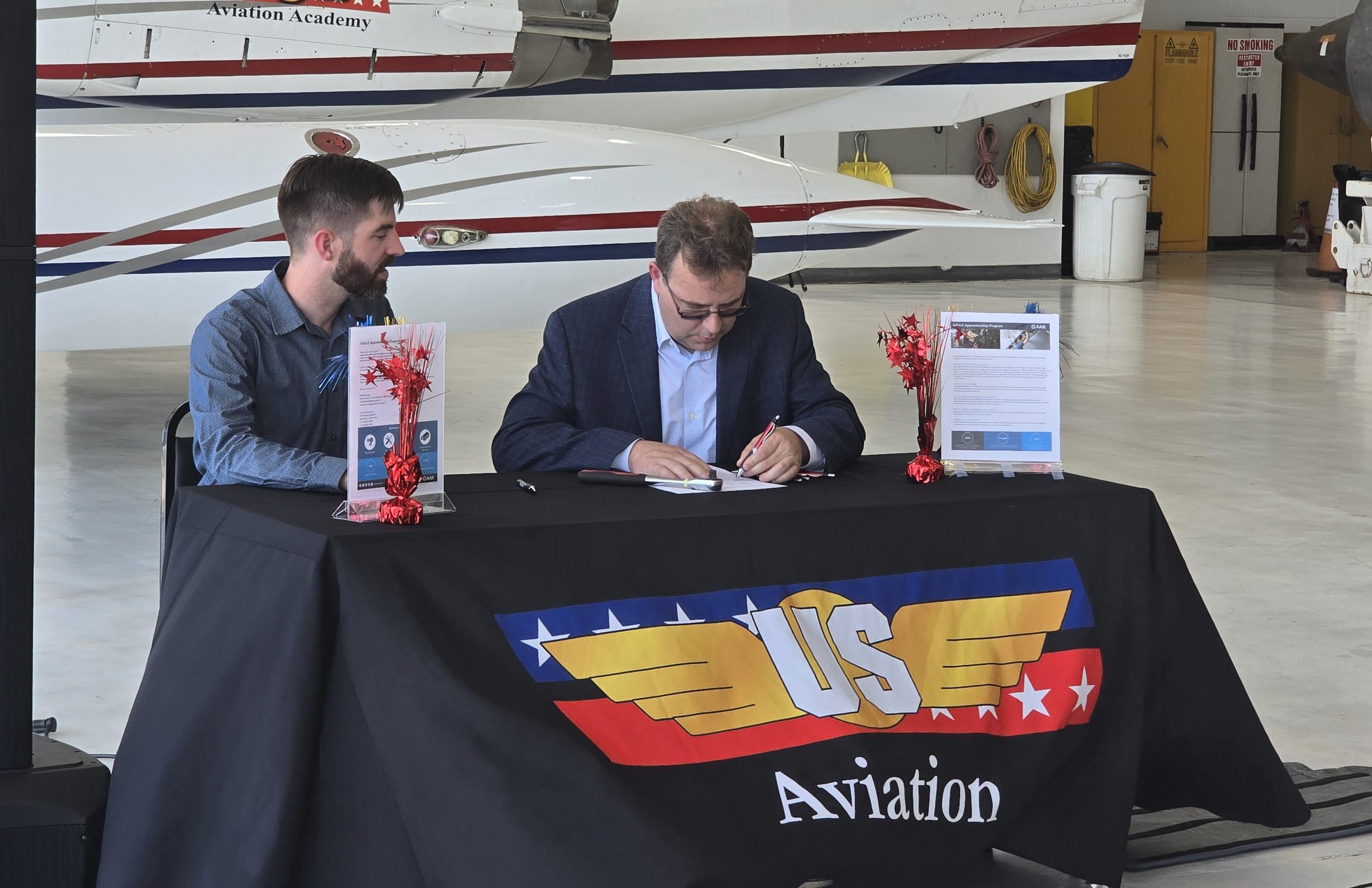 US Aviation Academy partnership signing ceremony where an executive in a suit signs an official document with a colleague watching inside the flight school hangar