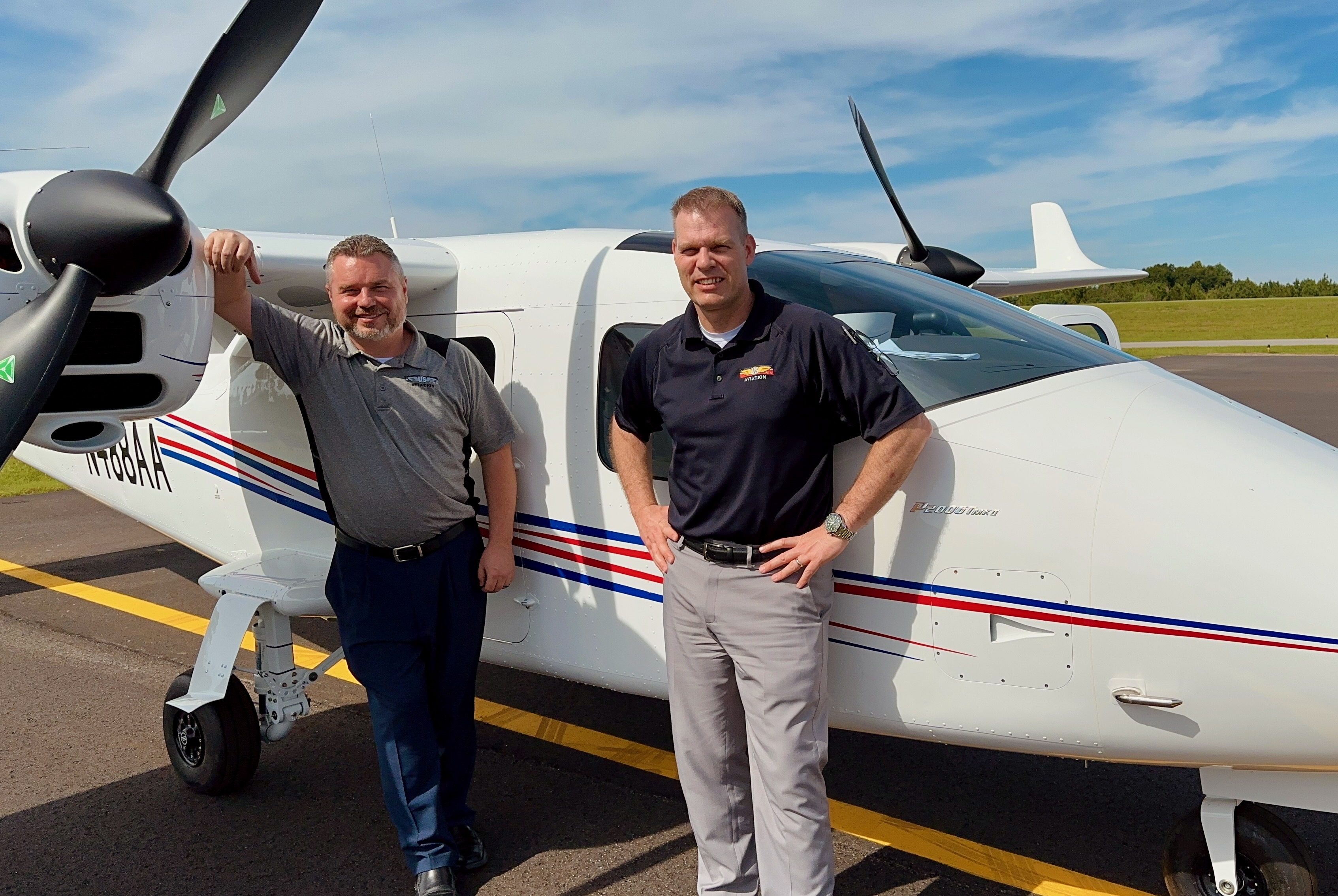 Two men standing beside a white twin-engine aircraft on the tarmac, smiling and posing near the propeller.